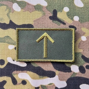 以下が含まれることがあります： 金色の矢印が上を向いている長方形の緑色のパッチ。金色の糸で縫われています。パッチは金色の縁取りがあり、迷彩柄の背景に配置されています。