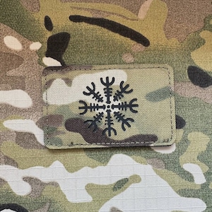 Könnte beinhalten: Ein schwarz-weißes Patch mit einem Symbol, das einer Windrose oder einem Schneeflocke ähnelt. Das Patch ist aus einem strukturierten Stoff gefertigt und hat einen schwarzen Rand. Das Patch befindet sich auf einem Tarnmuster-Hintergrund.