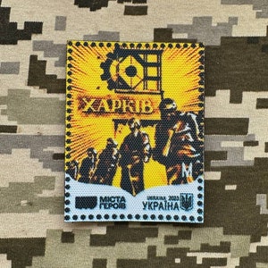 Puede incluir: Un parche amarillo y negro con una imagen estilizada de un engranaje y tres figuras. El texto "ХАРКІВ" es visible en el centro del parche. El texto "МІСТА ГЕРОЇВ" y "UKRAINA 2023 УКРАЇНА" es visible en la parte inferior del parche.