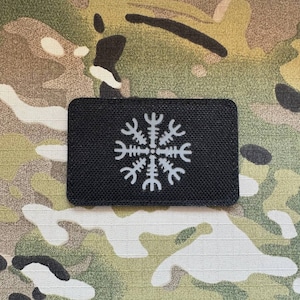 Puede incluir: Un parche negro con un símbolo blanco que se asemeja a un copo de nieve o una estrella de ocho puntas. El símbolo es una versión estilizada del Vegvisir, un símbolo direccional nórdico.