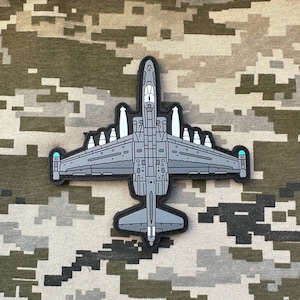 Peut inclure: Un écusson brodé gris et blanc représentant un avion militaire avec un design détaillé. L'écusson est sur un fond de tissu camouflage.