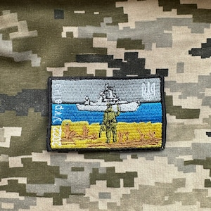 Könnte beinhalten: Ein gestickter Aufnäher in Gelb und Blau mit einem Schiff und einem Soldaten. Der Aufnäher zeigt den Text "Ehre der Ukraine" und das ukrainische Dreizack-Symbol.