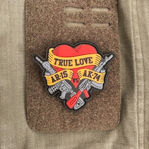 Könnte beinhalten: Ein schwarzer und grauer Patch mit einem roten Herzen und einem gelben Banner, auf dem "TRUE LOVE" steht, darunter zwei gekreuzte Waffen. Das Banner sagt "AR-15 VS AK-74".