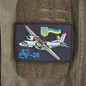 Könnte beinhalten: Ein blaues und weißes gesticktes Abzeichen mit einem Flugzeug und der ukrainischen Flagge. Das Flugzeug ist mit "An-26" beschriftet.