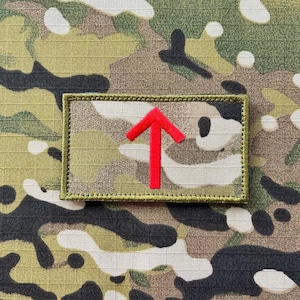 以下が含まれることがあります： 緑と茶色の迷彩生地のパッチに、上向きに指し示す赤い矢印が描かれている。