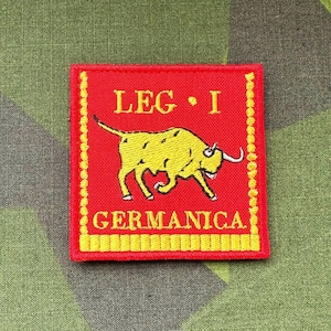 Puede incluir: Un parche bordado rojo y amarillo con el texto "LEG I GERMANICA" y un toro amarillo corriendo hacia la derecha.