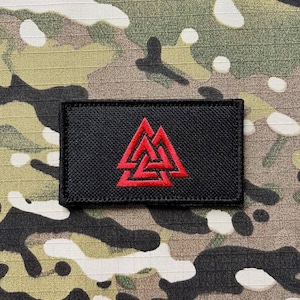 May include: A black patch with a red embroidered Valknut symbol. The patch is made of a textured fabric and has a white border.