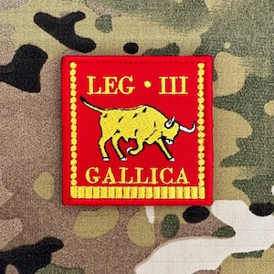 Puede incluir: Un parche rojo bordado con texto amarillo y una ilustración de toro amarillo. El texto dice "LEG III GALLICA". El parche cuadrado tiene un borde amarillo y está sobre un fondo de camuflaje.