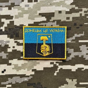 Könnte beinhalten: Ein gelber gestickter Aufnäher mit einer blau-schwarzen ukrainischen Flagge und einem Hammer-und-Sichel-Symbol. Der Text "ДОНЕЦЬК ЦЕ УКРАЇНА!" ist in Gelb auf dem Aufnäher geschrieben.