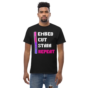 May include: Black t-shirt with the text "EMBED CUT STAIN REPEAT" in white and pink. A vertical design with pink, purple, and blue colors is on the left side of the text. The shirt is made of a soft material.