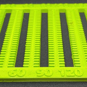 Può includere: Uno strumento in plastica verde neon brillante con fessure verticali e marcature numerate. I numeri 30, 60, 90, 120 e 150 sono visibili. Lo strumento sembra essere una guida di misurazione o spaziatura.