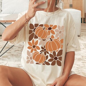 May include: Cream-colored t-shirt featuring a fall-themed design. The graphic includes three orange pumpkins and various brown and tan flowers. The shirt is a relaxed fit, suitable for casual wear.