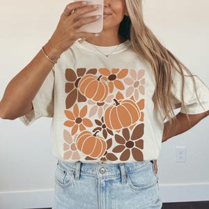 May include: Cream-colored t-shirt featuring a fall-themed design. The graphic includes orange pumpkins and flowers in shades of brown and beige. The shirt is paired with light blue denim shorts.