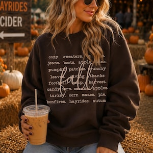 May include: A dark chocolate brown long-sleeve sweatshirt featuring white text that lists fall-themed words including cozy sweaters, jeans, boots, apple cider, pumpkin patches, crunchy leaves, giving thanks, football, soup, harvest, candy corn, caramel apples, turkey, corn mazes, pie, bonfires, hayrides, and autumn.