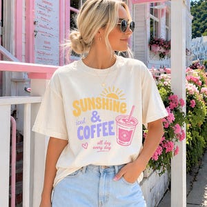 Puede incluir: Camiseta color crema con el texto "Sunshine, iced & coffee, all day every day" en amarillo, morado y rosa. La camiseta presenta un gr&aacute;fico de una bebida de caf&eacute;. La modelo lleva unos shorts vaqueros azul claro.