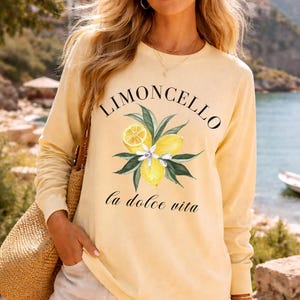 May include: A light yellow long-sleeve shirt with the word "Limoncello" above a watercolor illustration of lemons and leaves. The phrase "La dolce vita" is printed below the illustration. The shirt is worn with shorts and a straw tote bag.