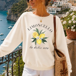 May include: Ivory-colored sweatshirt with the word "LIMONCELLO" above a lemon and leaf graphic. Below the graphic is the phrase "la dolce vita". The sweatshirt is worn with white shorts and a woven tote bag.