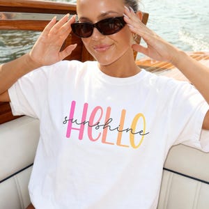 May include: White t-shirt with the text "HELLO sunshine" in pink, orange, and yellow. The model is wearing sunglasses and is on a boat.