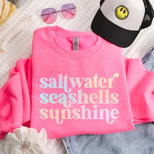 May include: A bright pink sweatshirt with the words "saltwater seashells sunshine" in pastel colors. The image also includes a black and white trucker hat with a yellow smiley face, pink sunglasses, and blue jeans.