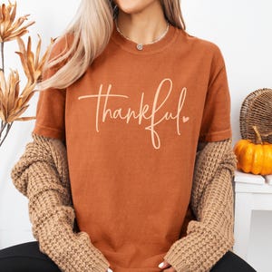 May include: A yam-colored t-shirt with the word "thankful" in a cursive font. The shirt is paired with a tan sweater and black pants. A small pumpkin and decorative elements are in the background.