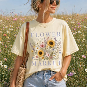 May include: A pale yellow short-sleeve t-shirt featuring a graphic print of various colorful wildflowers, including sunflowers, with the text "FLOWER MARKET" in blue lettering. The shirt is paired with light blue denim jeans and a woven bag.