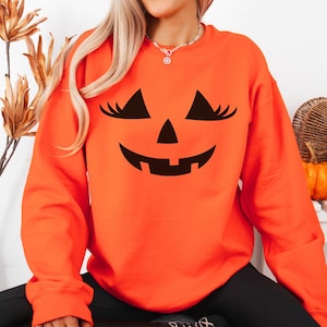 May include: A bright orange long-sleeve sweatshirt featuring a black graphic print of a jack-o'-lantern face with eyelashes on the front. The design includes triangular eyes with long lashes, a triangular nose, and a wide, toothy grin.