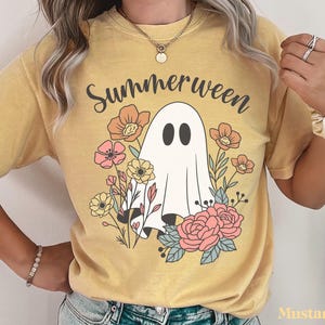 May include: Mustard-colored t-shirt with the word "Summerween" above a cartoon ghost surrounded by flowers. The ghost has two black oval eyes and is draped in a white sheet. The flowers are in shades of pink, orange, and yellow.