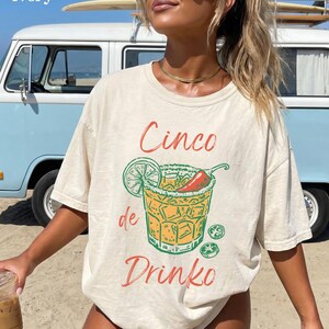 May include: Ivory-colored t-shirt with a Cinco de Drinko graphic. The design features a cocktail with a lime, chili pepper, and ice cubes. The text "Cinco de Drinko" is in red and green. The shirt is worn on a beach.