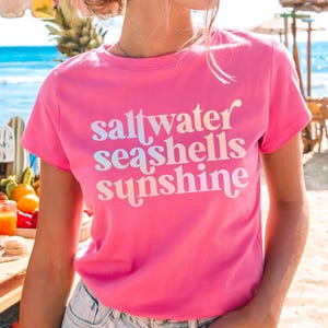 May include: A bright pink t-shirt with the words "saltwater seashells sunshine" in a white and light blue font. The shirt is short-sleeved and has a round neckline. The text "Charity Pink" is at the bottom right corner of the image.