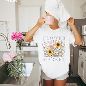 May include: A white short-sleeved t-shirt featuring a graphic print of a colorful wildflower bouquet with sunflowers and the text "FLOWER MARKET" in purple lettering. The shirt is worn with grey shorts and a white towel wrapped around the head.