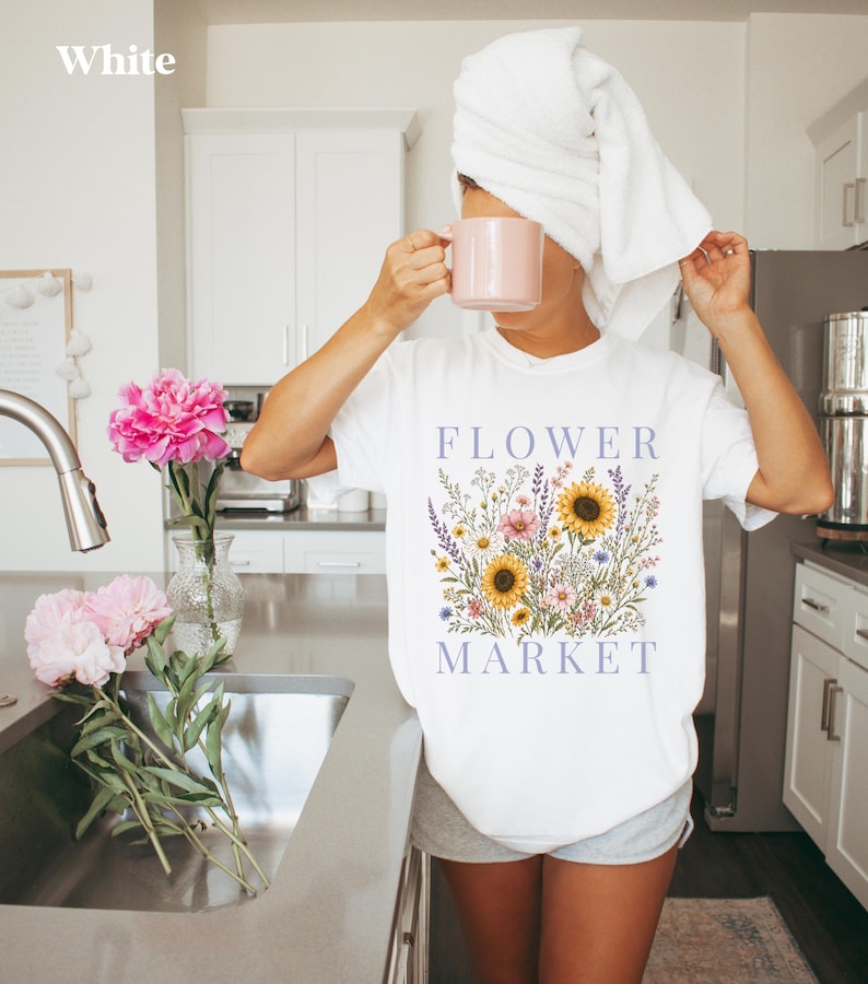 May include: A white short-sleeved t-shirt featuring a graphic print of a colorful wildflower bouquet with sunflowers and the text "FLOWER MARKET" in purple lettering. The shirt is worn with grey shorts and a white towel wrapped around the head.