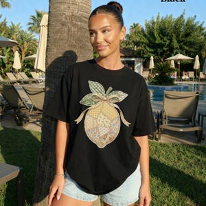 May include: Black t-shirt with a patchwork lemon design, green leaves, and a bow. The word "Black" is printed in the upper right corner. The shirt is paired with light blue denim shorts.