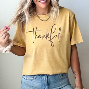 May include: Mustard-colored t-shirt with the word "thankful" in a brown script font. A small pink heart is next to the word. The shirt is worn with blue jeans.