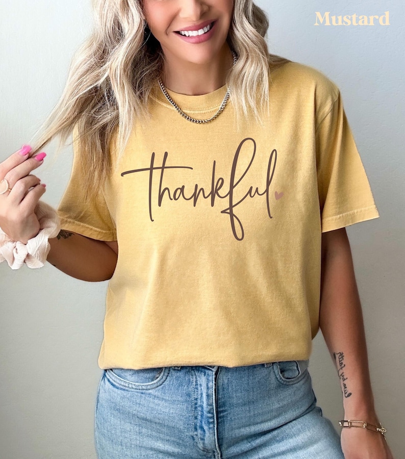 May include: Mustard-colored t-shirt with the word "thankful" in a brown script font. A small pink heart is next to the word. The shirt is worn with blue jeans.