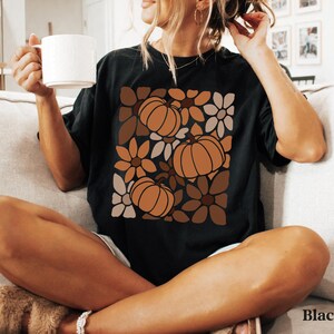 May include: Black t-shirt featuring a pumpkin and floral design in shades of brown and orange. The shirt has a relaxed fit and short sleeves. The word "Black" is printed on the bottom right corner of the image.