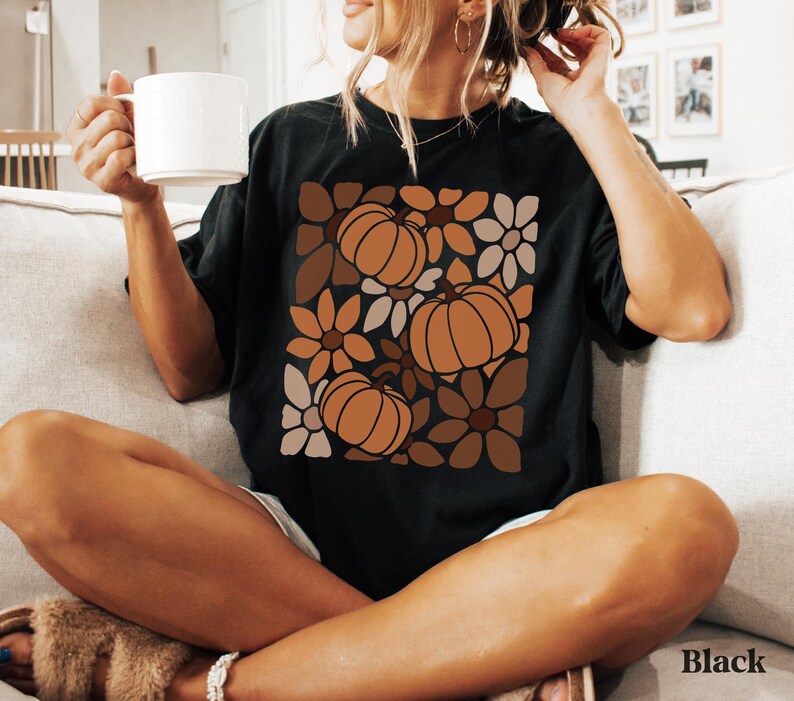 May include: Black t-shirt featuring a pumpkin and floral design in shades of brown and orange. The shirt has a relaxed fit and short sleeves. The word "Black" is printed on the bottom right corner of the image.