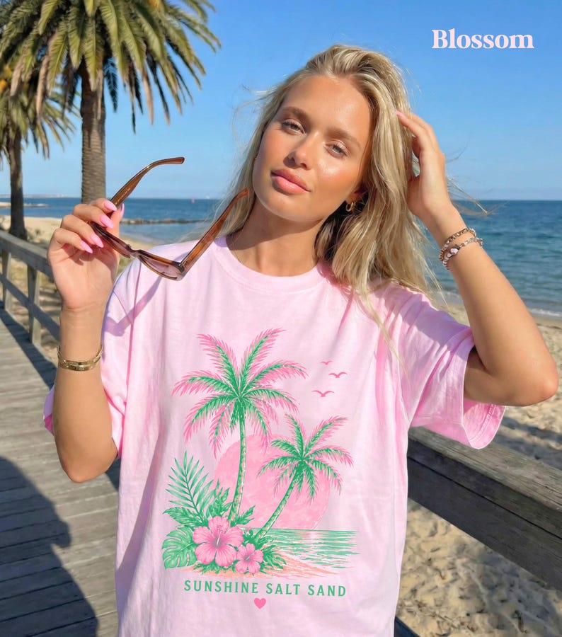 May include: Pale pink t-shirt with a tropical design. The graphic includes palm trees, a pink sun, and the text "Sunshine Salt Sand." The design also features pink hibiscus flowers and green leaves. The shirt is a light pink colour.