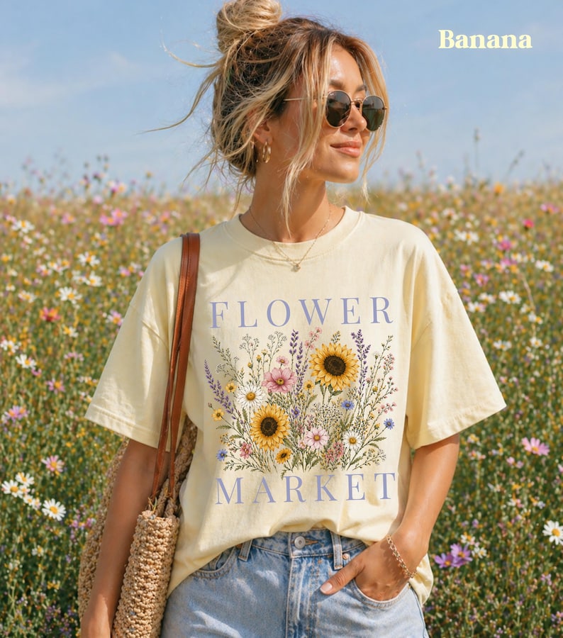 May include: A pale yellow short-sleeve t-shirt featuring a graphic print of various colorful wildflowers, including sunflowers, with the text "FLOWER MARKET" in blue lettering. The shirt is paired with light blue denim jeans and a woven bag.