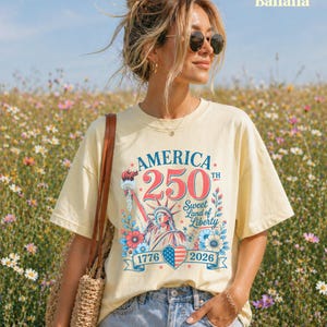 K&ouml;nnte beinhalten: Hellgelbes T-Shirt mit einem patriotischen Design zum 250. Jahrestag Amerikas. Die Grafik zeigt die Freiheitsstatue, florale Elemente und den Text "America 250th Sweet Land of Liberty 1776-2026."