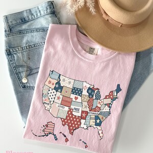 May include: Pink t-shirt with a patchwork design of the United States map. The map is made up of different colored squares with stars and stripes. The words "Brave", "Loved", "Free", and "Victorious" are printed on the map.