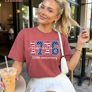 May include: A crimson t-shirt with the text "1776 america 250th anniversary" in a patriotic design. The shirt features red and blue stars and stripes. The model is wearing a white skirt and holding a drink.