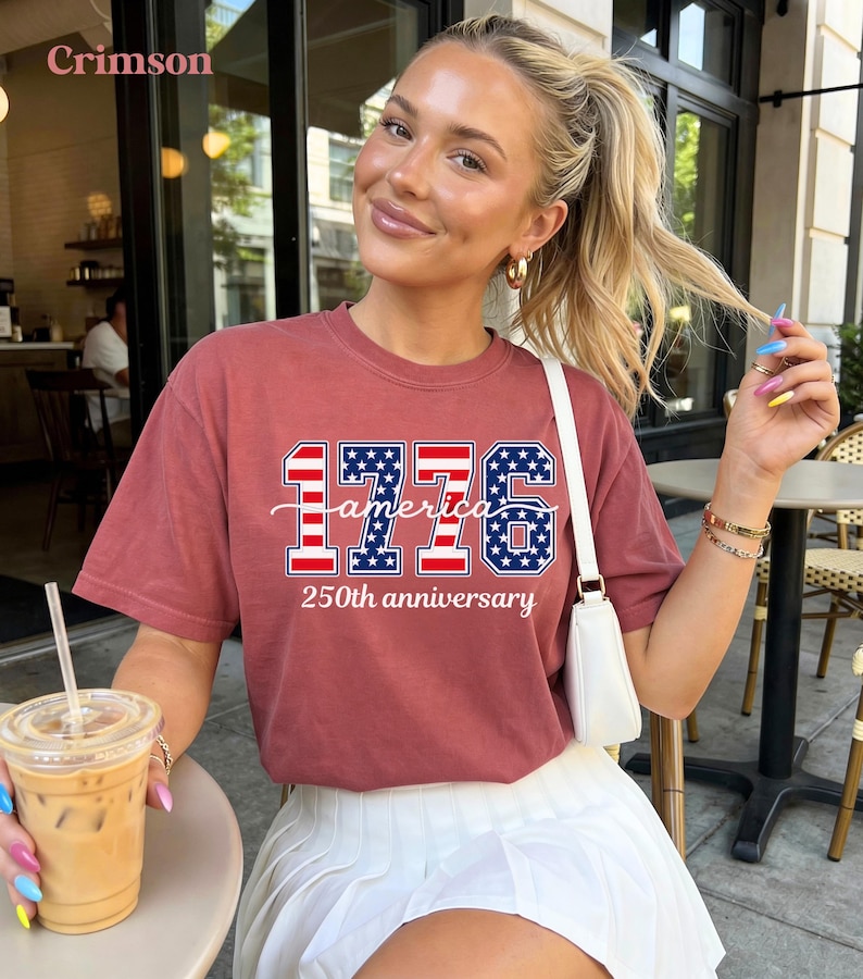 May include: A crimson t-shirt with the text "1776 america 250th anniversary" in a patriotic design. The shirt features red and blue stars and stripes. The model is wearing a white skirt and holding a drink.
