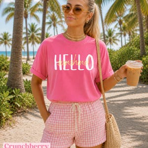 May include: A vibrant pink t-shirt with the text "Hello Sunshine" in white and yellow. The model is wearing pink and white gingham shorts and sunglasses. A straw bag is over the shoulder. The background includes palm trees and a beach setting.