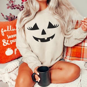 May include: A sand-coloured crewneck sweatshirt featuring a black graphic of a jack-o'-lantern face with long eyelashes. The word Sand is printed in the top right corner. The garment has long sleeves and a ribbed neckline, cuffs, and hem.