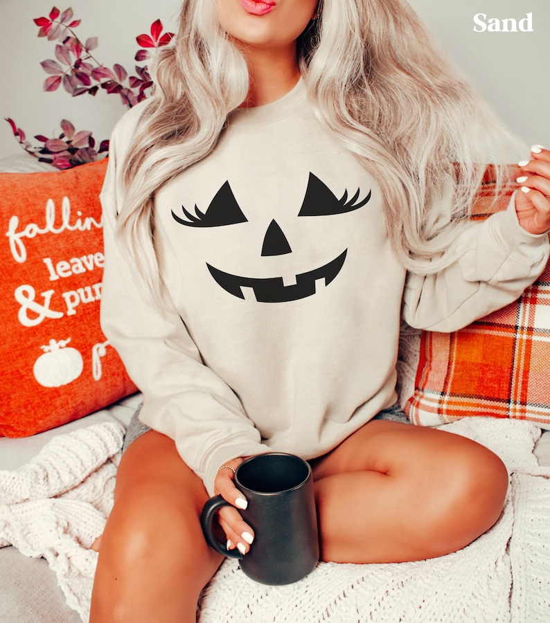May include: A sand-coloured crewneck sweatshirt featuring a black graphic of a jack-o'-lantern face with long eyelashes. The word Sand is printed in the top right corner. The garment has long sleeves and a ribbed neckline, cuffs, and hem.