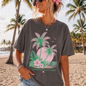 May include: Dark grey t-shirt with a tropical graphic of pink and green palm trees, a pink sun, and the text "Sunshine Salt Sand." The shirt is worn on a beach with palm trees in the background.