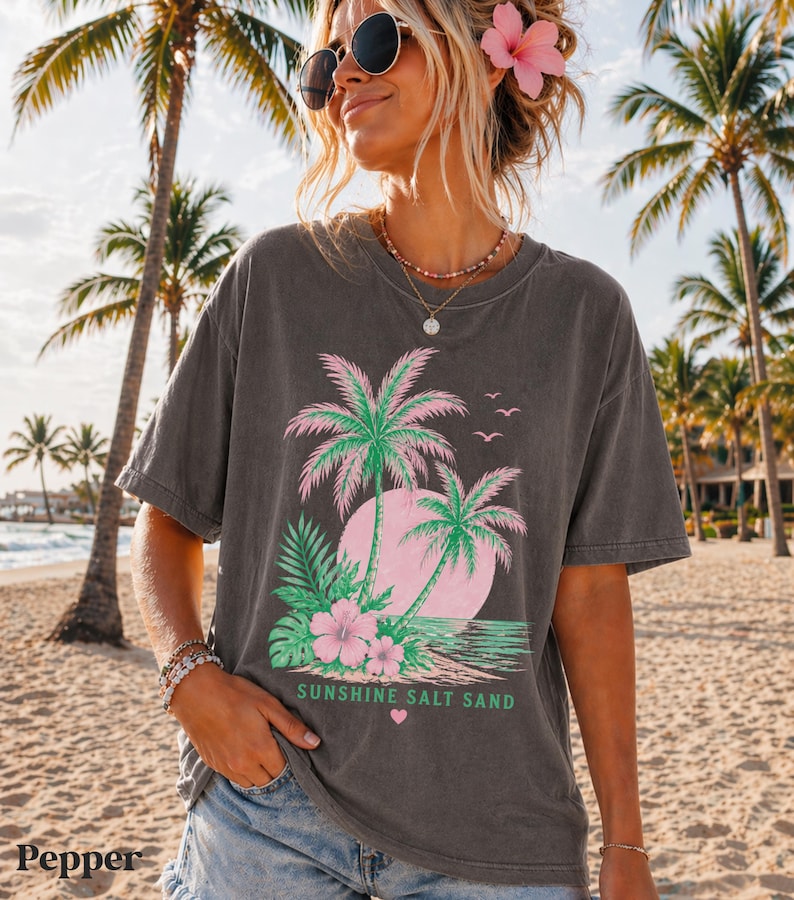 May include: Dark grey t-shirt with a tropical graphic of pink and green palm trees, a pink sun, and the text "Sunshine Salt Sand." The shirt is worn on a beach with palm trees in the background.
