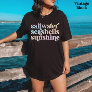 May include: Black t-shirt with the words "saltwater seashells sunshine" in a gradient font. The shirt is a vintage black color and has a relaxed fit. The background shows a wooden pier and the ocean.