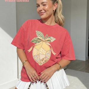 May include: A red t-shirt with a patchwork lemon design. The lemon features a yellow and floral pattern with green leaves and a bow. The word "Watermelon" is printed at the top of the image.