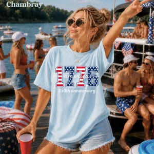 May include: A light blue t-shirt featuring the text "1776 America 250th anniversary" in red, white, and blue. The shirt is paired with denim shorts. The image is taken outdoors near a lake with people and boats in the background.